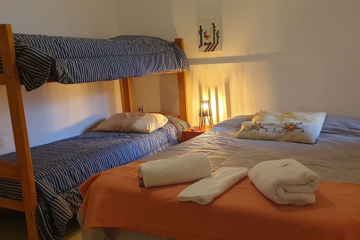 A room with two beds, one with a bunk and one with a single mattress. There are pillows and a blanket on the beds, and a lamp on the nightstand.