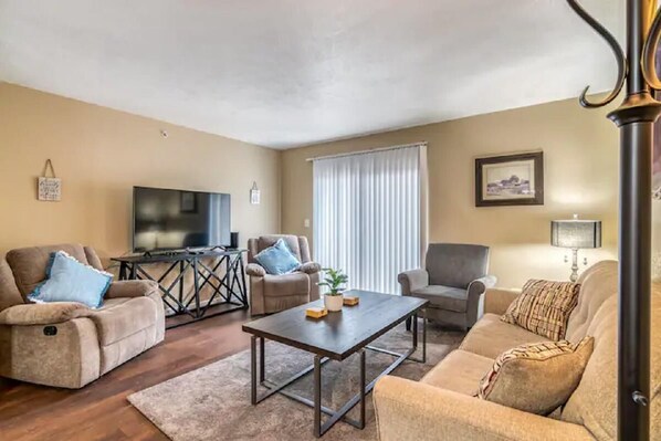 A living room with a sofa, armchairs, a coffee table, a TV, and a window with curtains.