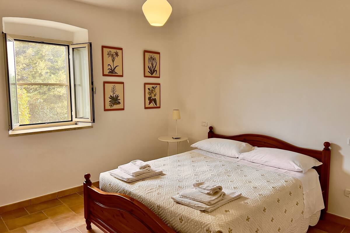 A bedroom with a wooden bed, white bedding, a small bedside table, a lamp, and four framed floral pictures on the wall.