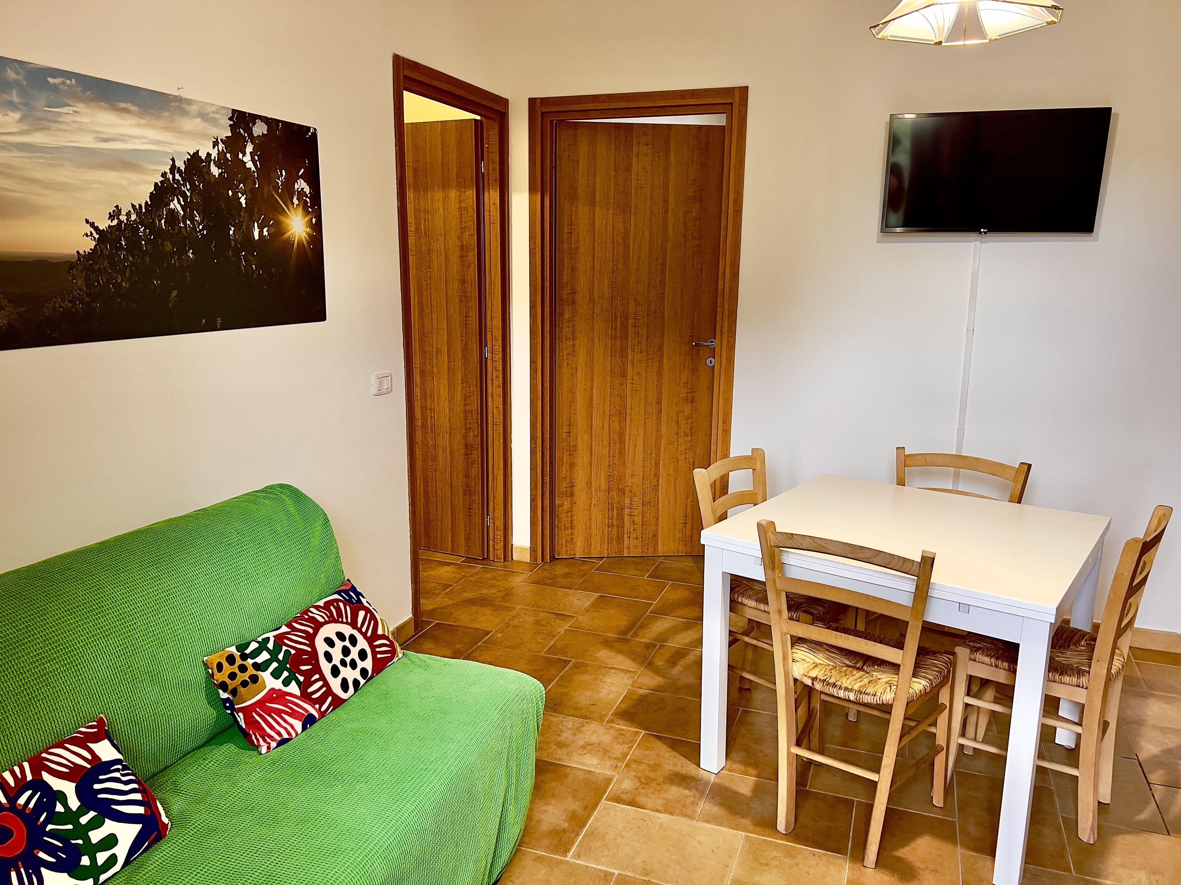 Een kamer met een groene bank, een witte tafel met stoelen, een houten deur en een televisie aan de muur.