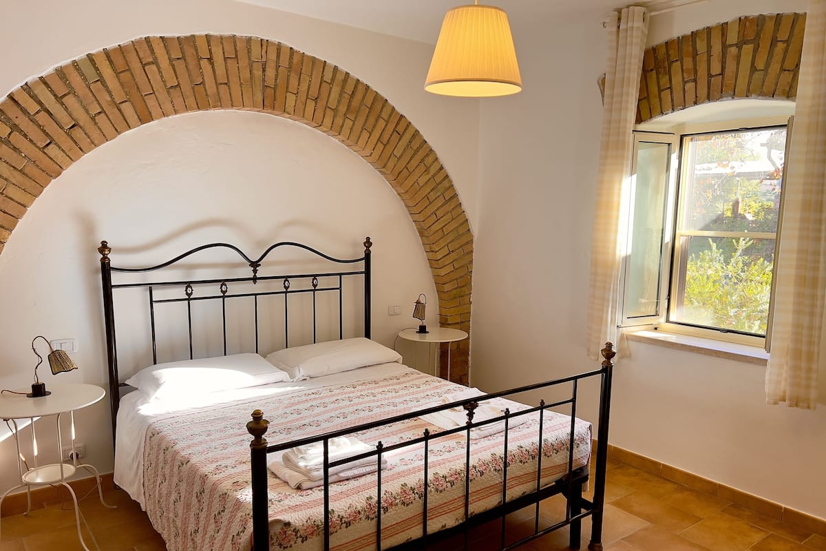 A bedroom with a brick arch, a bed with a floral bedspread, a bedside table with a lamp, and a window with a view of greenery.