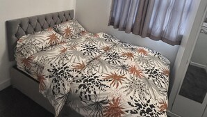 Una cama individual con cabecera capitonada, colcha estampada con motivos florales y ventana con cortina.