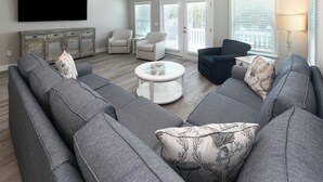 A modern living room with a grey sectional sofa, two armchairs, a round coffee table, and a flat-screen TV mounted on the wall.