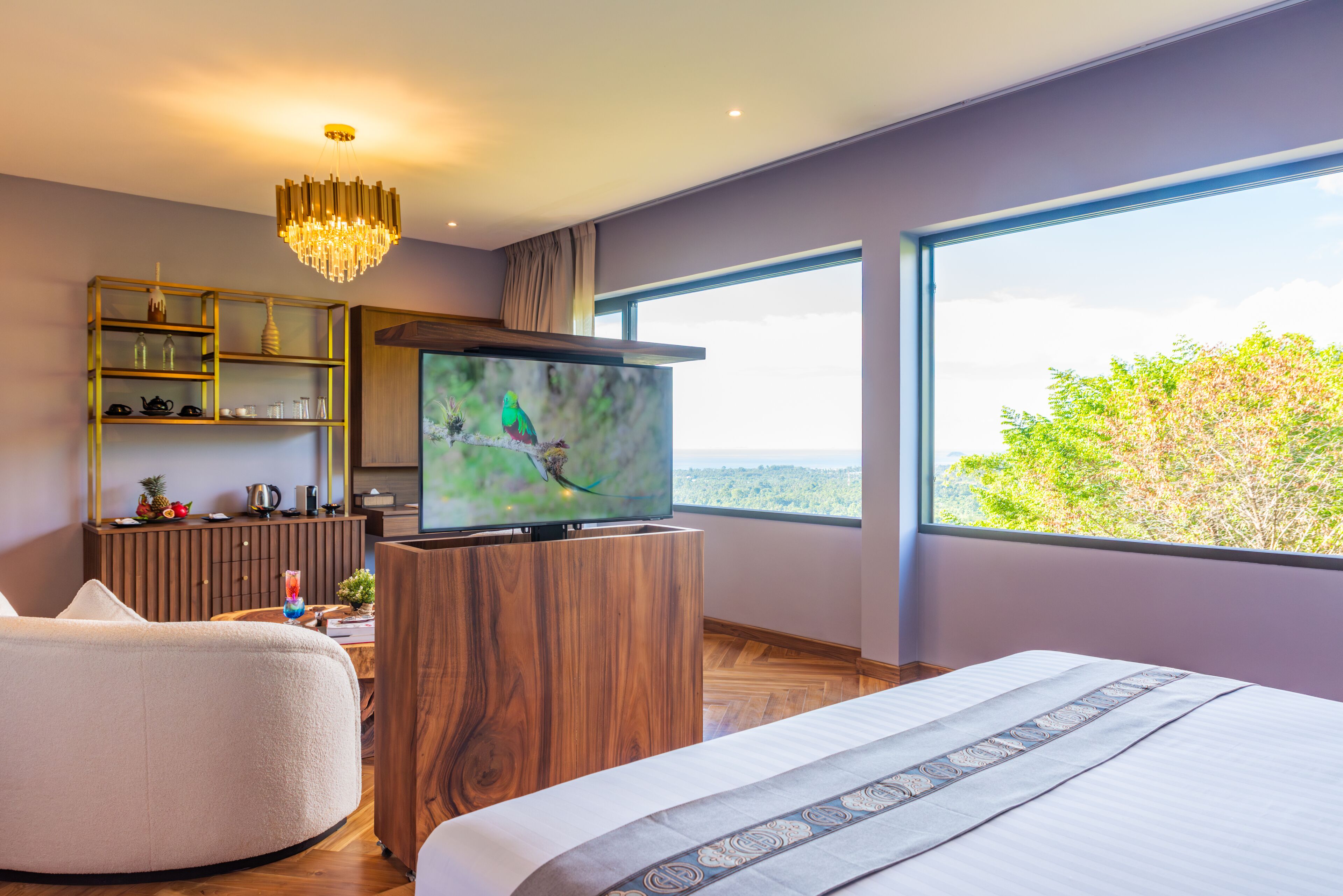A modern bedroom with a large bed, a television on a wooden stand, a seating area, and a large window offering a view of the outdoors.