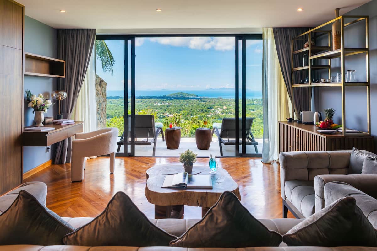 Un vaste espace de vie agrémenté d’une grande fenêtre offrant une vue sur un paysage luxuriant. On y trouve des sièges confortables, une table basse et une bibliothèque.