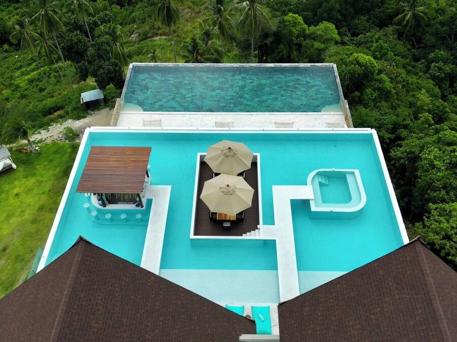 An aerial view of a resort with a swimming pool, lounge area, and surrounding lush greenery.