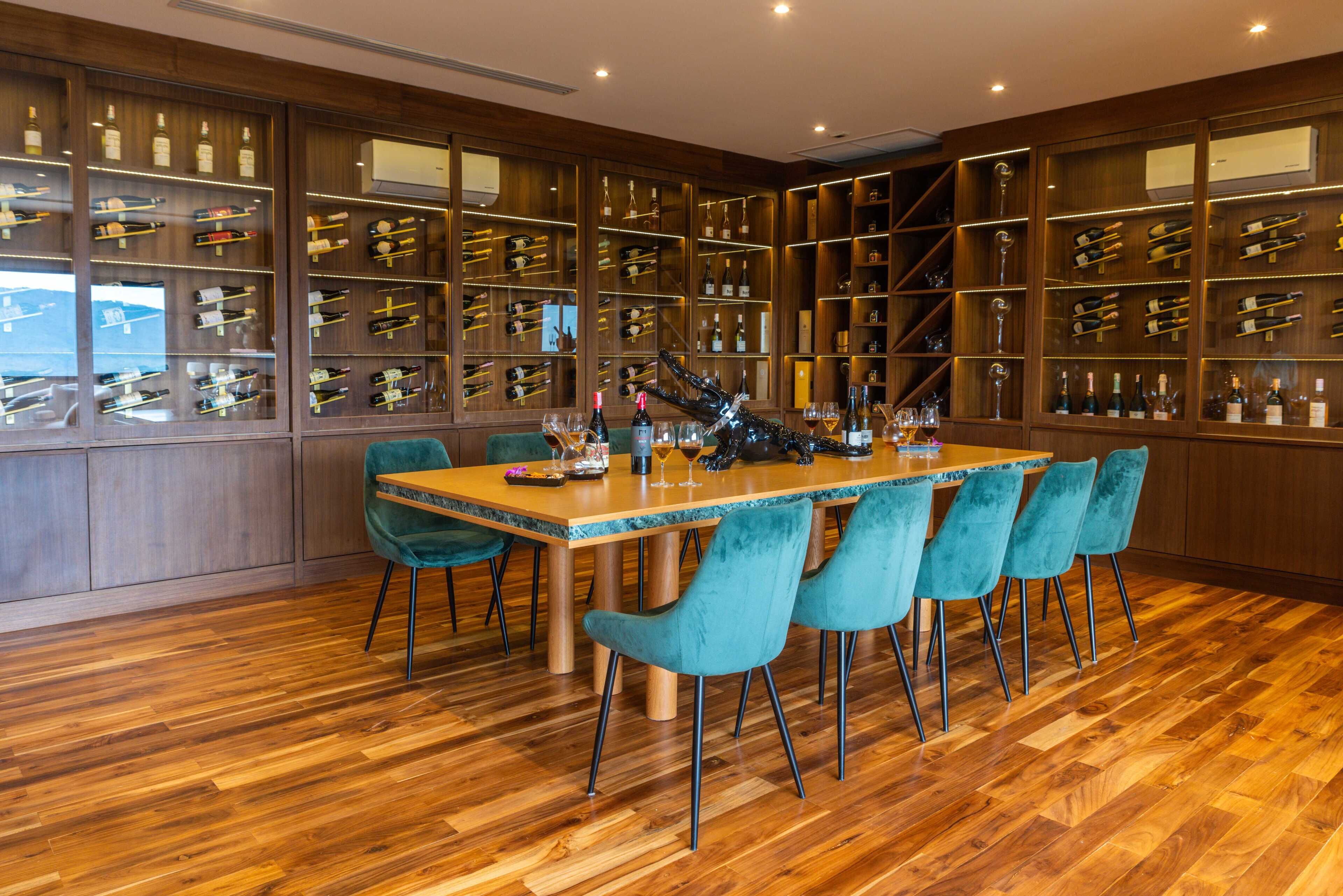 A dining area with a wooden table and teal chairs, surrounded by wine racks with bottles and glasses.