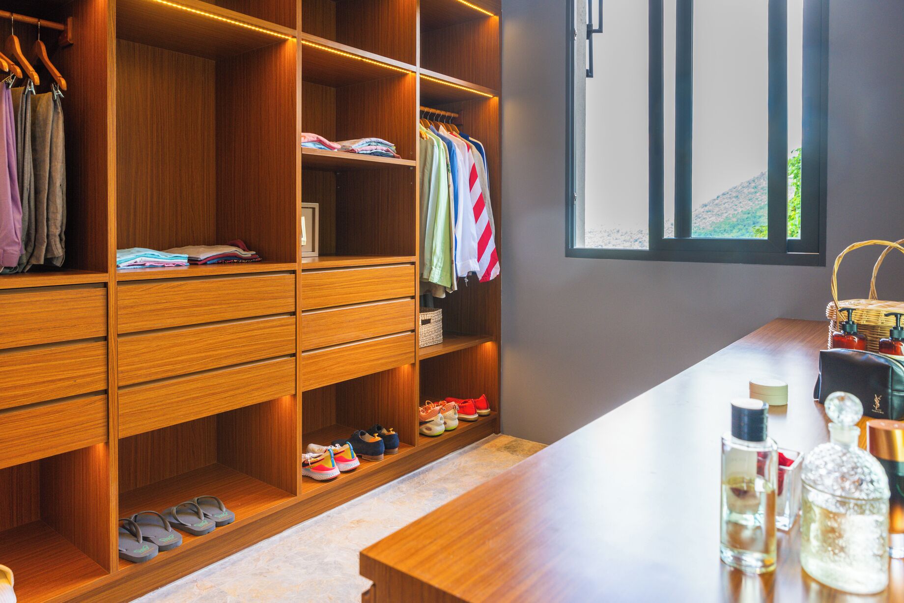 A well-organized closet with multiple shelves, drawers, and hanging space, a window with a view, and a wooden counter with bottles.
