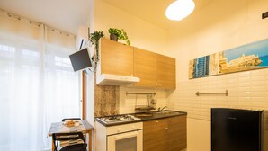 Een kleine keuken met een gasfornuis, oven en een zwarte koelkast. Er staat een klein tafeltje met een schaal eten en een plant op het aanrecht.