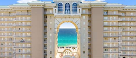A hotel with a central archway leading to a beach view, multiple balconies, and a symmetrical facade.