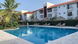 Une piscine entourée de chaises longues et d’ombrelles en feuilles de palmier, avec des palmiers tout autour et un immeuble à plusieurs étages doté de balcons.