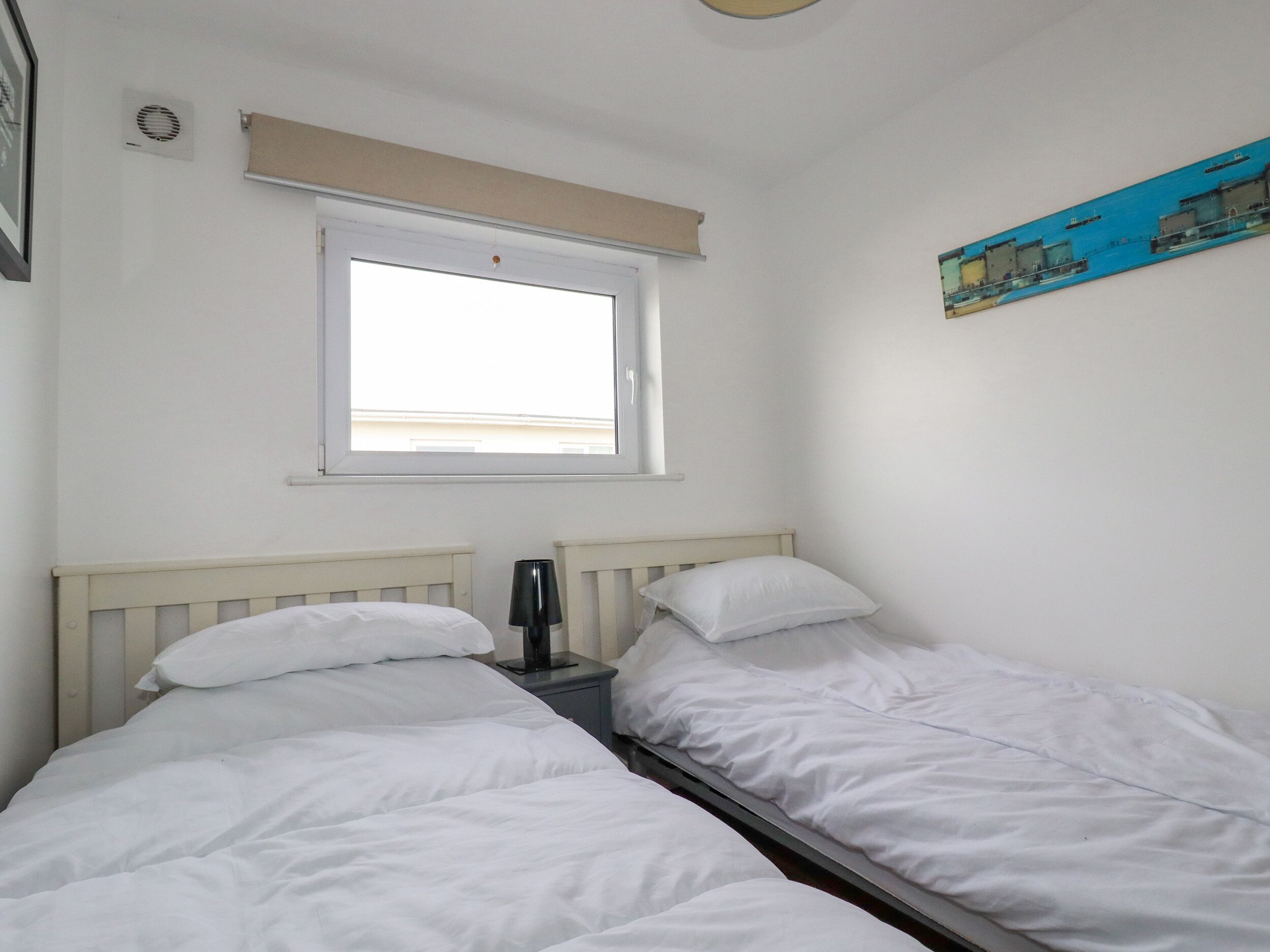 Ein Zimmer mit zwei Einzelbetten, einem Fenster mit Blick auf ein Gebäude, einem kleinen Tisch mit Lampe und einem an der Wand hängenden Bild.