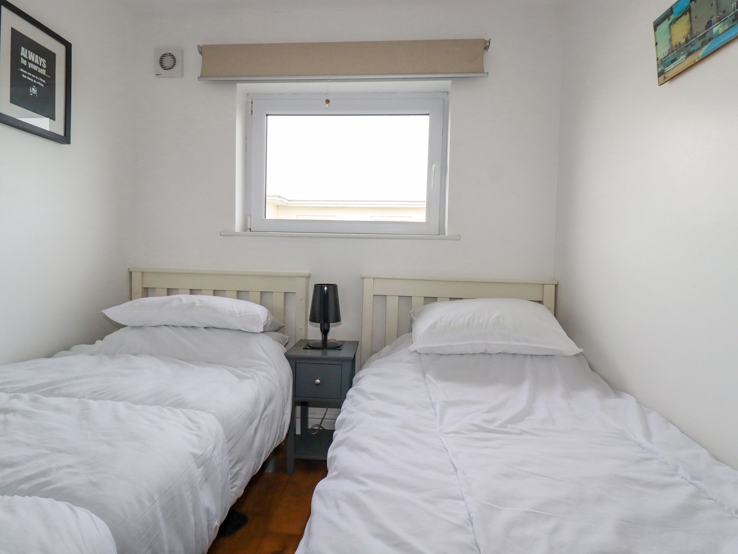 A bedroom with two single beds, a nightstand with a lamp, a window, and a framed quote on the wall.