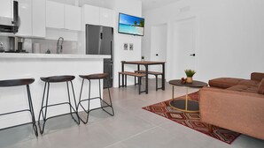 Cozinha moderna com armários brancos, geladeira de aço inoxidável e um espaço de bar com banquetas. Há uma mesa de jantar com bancos e uma pequena mesa lateral com uma planta. Um sofá marrom está na sala de estar, e uma TV está montada na parede.