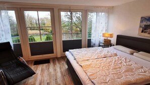 A hotel room with a bed, a chair, a lamp, and large windows with a view of greenery. - Charmante Hausvilla in Bojendorf (Bojendorf)