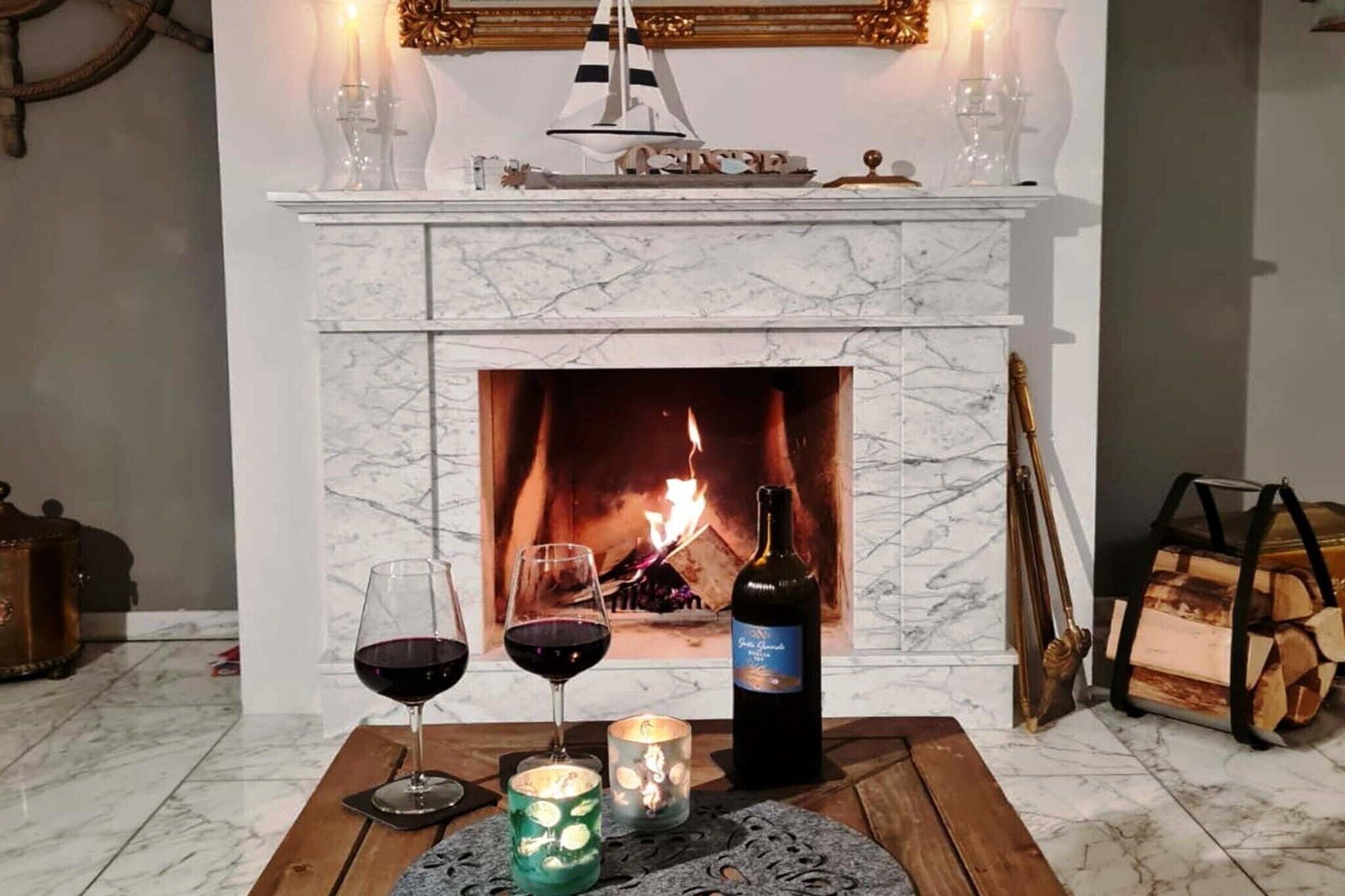 A fireplace with a lit fire, two wine glasses, a bottle of wine, and decorative items on a wooden table.