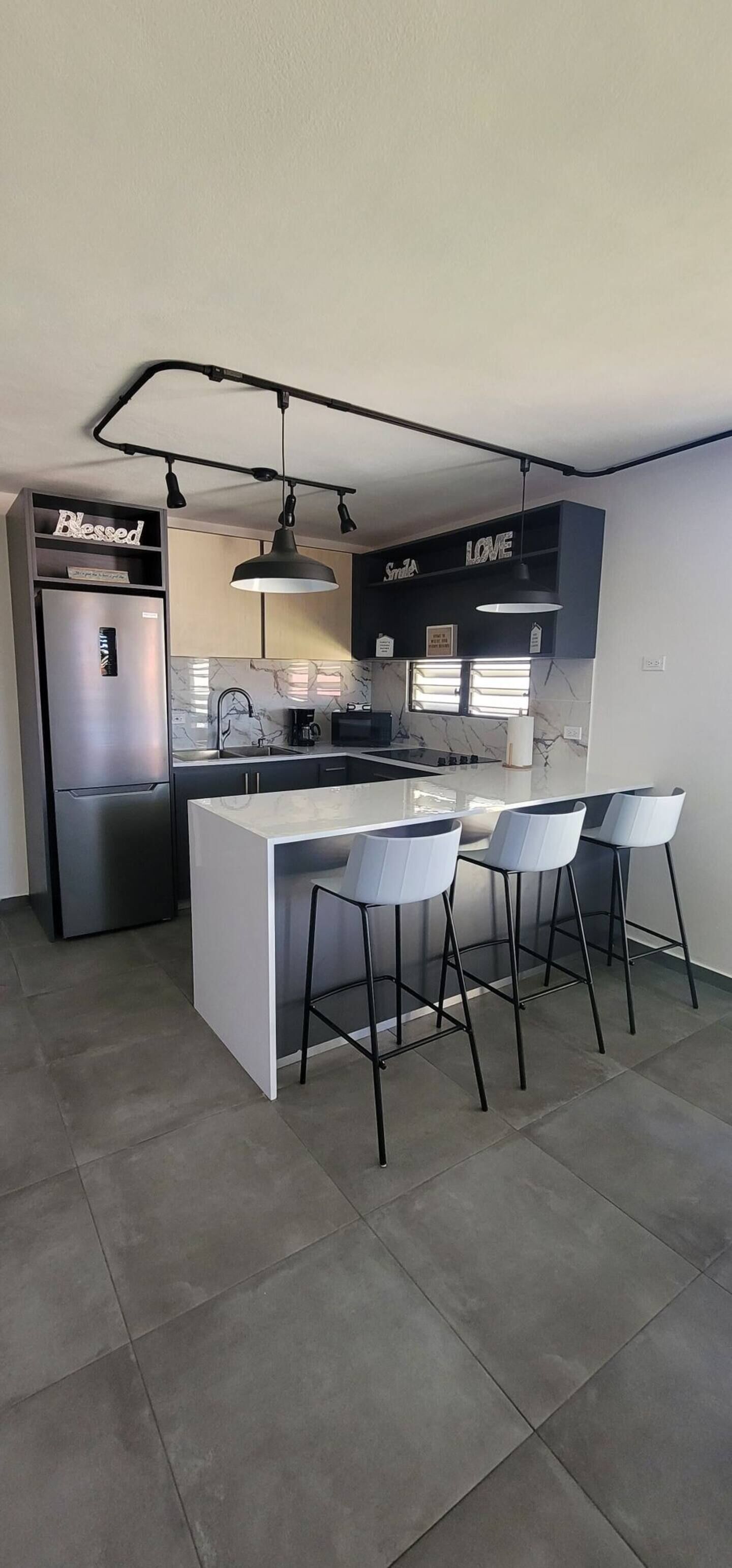 Una cocina moderna con una isla integrada, taburetes de bar, un refrigerador de acero inoxidable y una lámpara colgante.