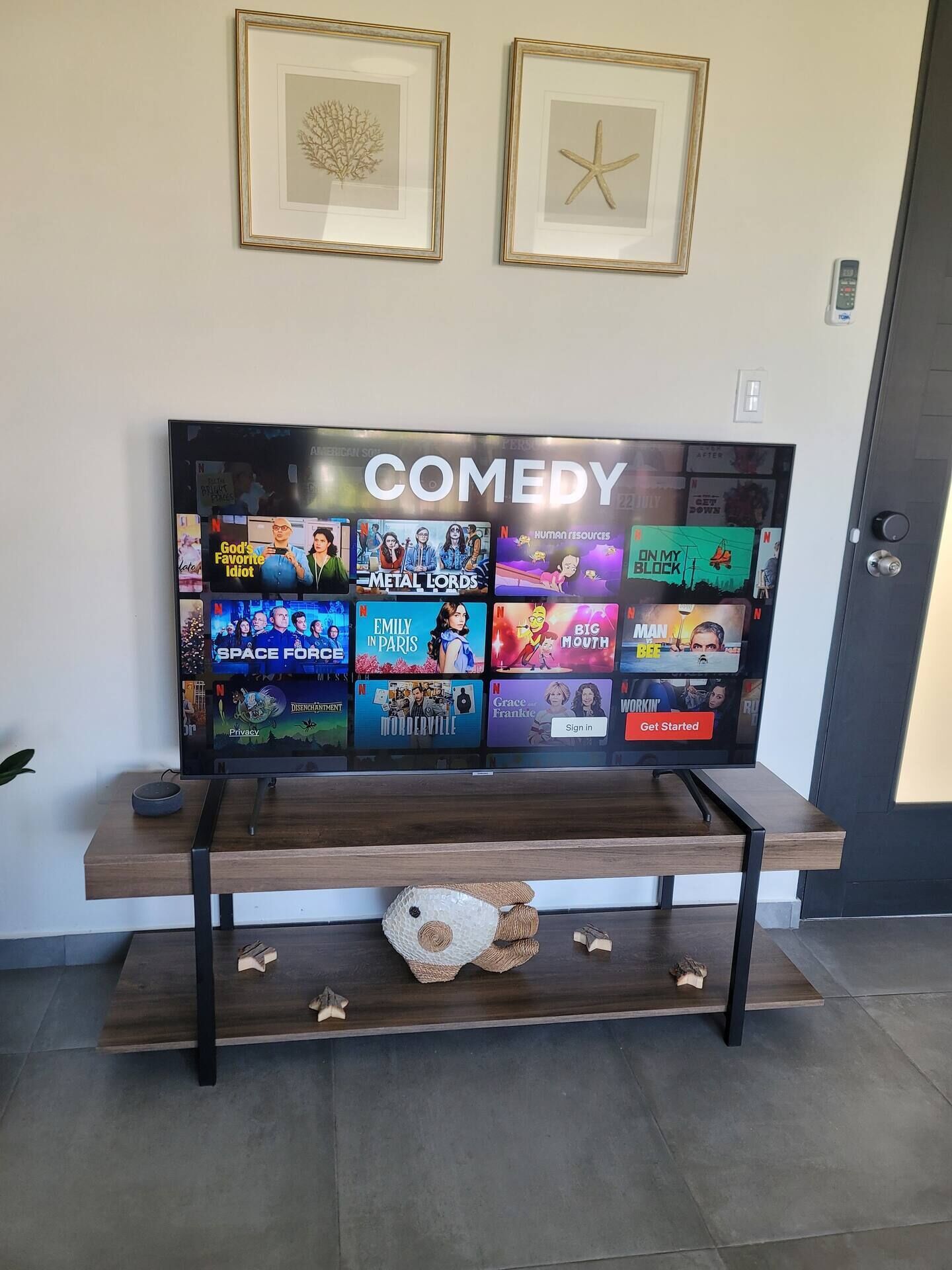 A flat-screen TV mounted on a wall with a 'COMEDY' label above it, displaying various Netflix shows.