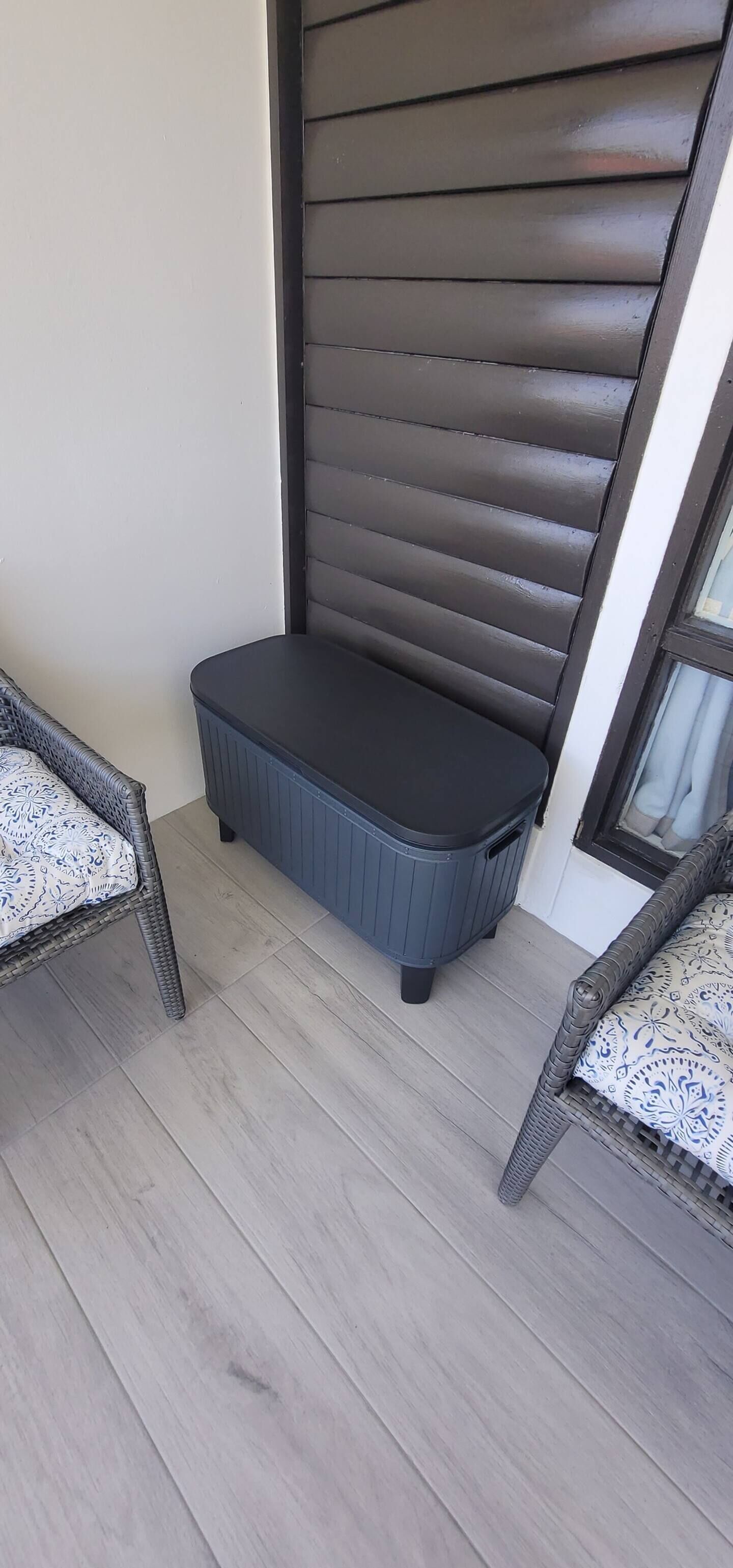 A small balcony with a dark-colored bench, two wicker chairs, and a light-colored wooden floor.