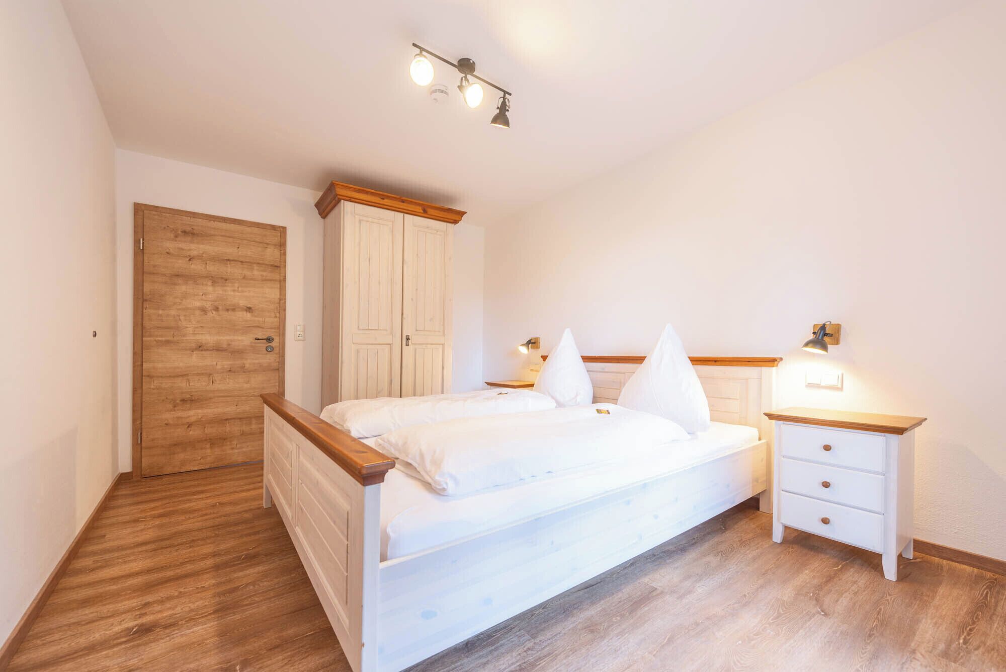 A bedroom with a wooden floor, a bed with white bedding, a nightstand with a lamp, and a wardrobe.