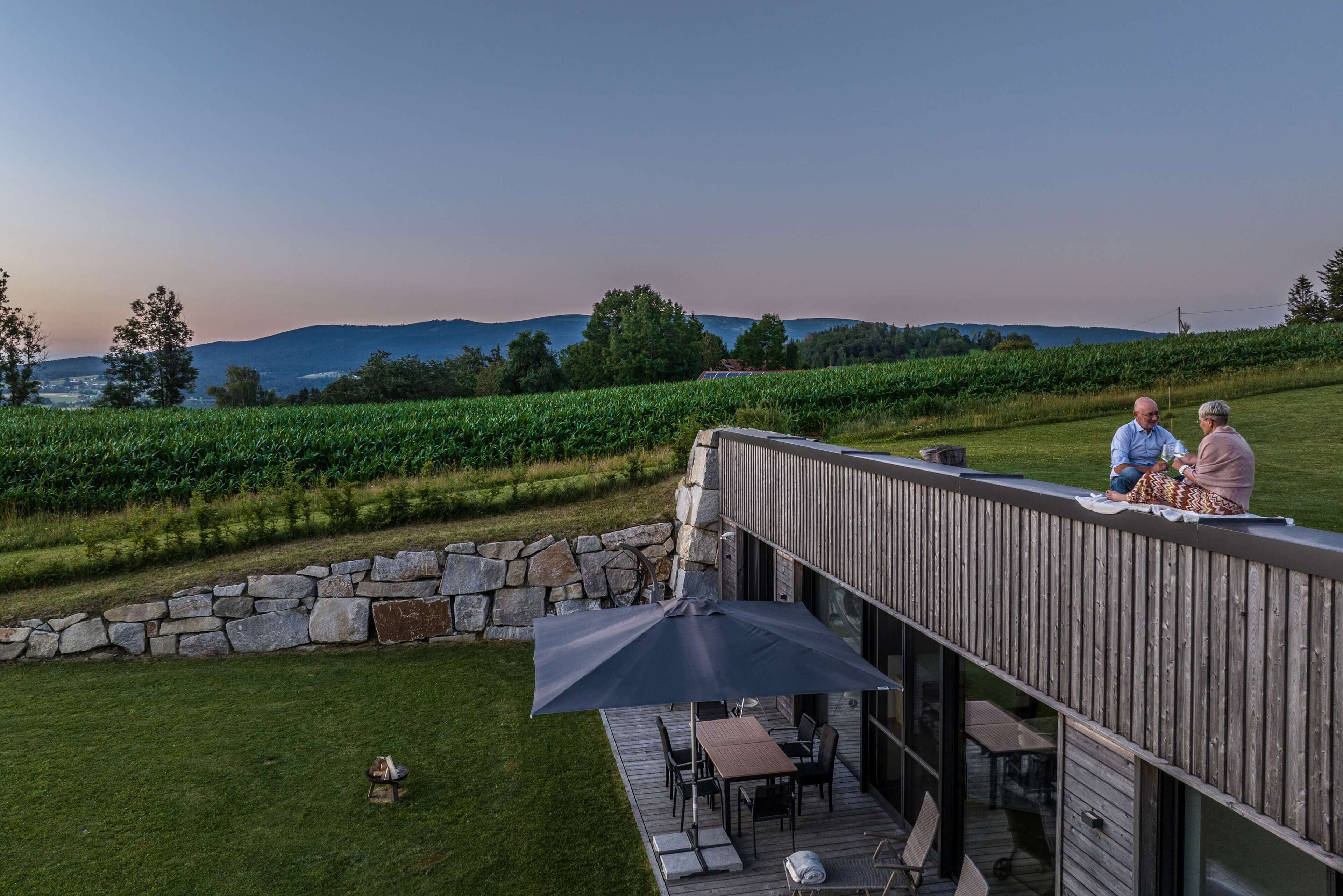 Eine Holzterrasse mit Gartenmöbeln, eine Steinmauer und Blick auf einen Weinberg und Hügel.