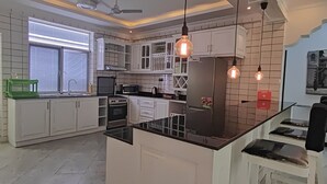 Una cocina moderna con armarios blancos, una isla central, iluminación colgante y un ventanal con persianas.