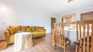 A modern living room with a yellow sofa, a round wooden coffee table, and a dining area with wooden chairs and a white tablecloth.