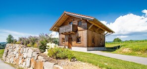 A wooden chalet with a balcony, stone wall, and a grassy landscape.
