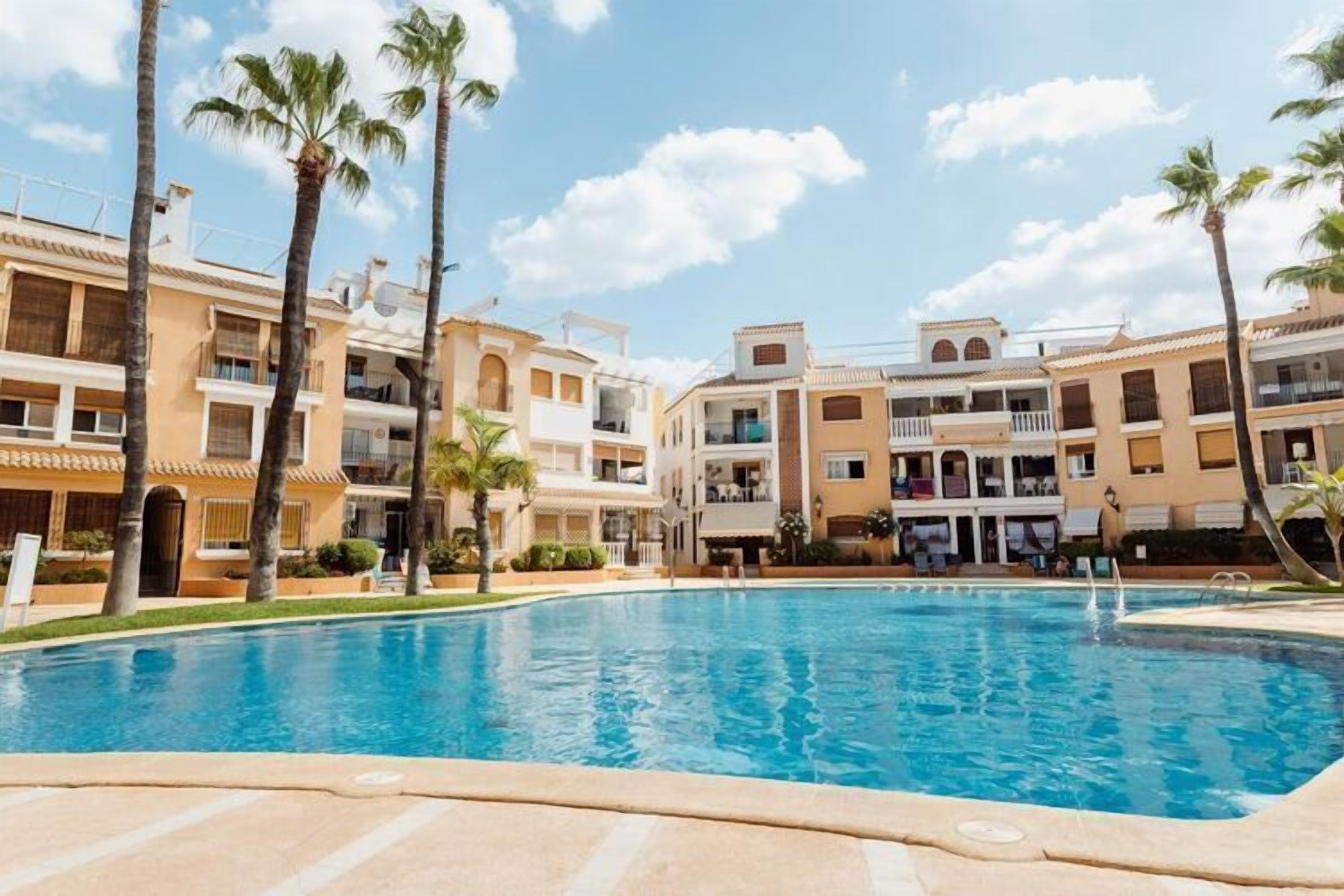 A swimming pool surrounded by multi-story buildings with balconies, palm trees, and a clear sky.