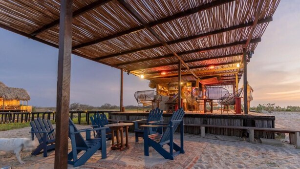 A beachside bar area with wooden chairs and tables, a thatched roof, and a view of the ocean. - Casa Solea (Manaure)