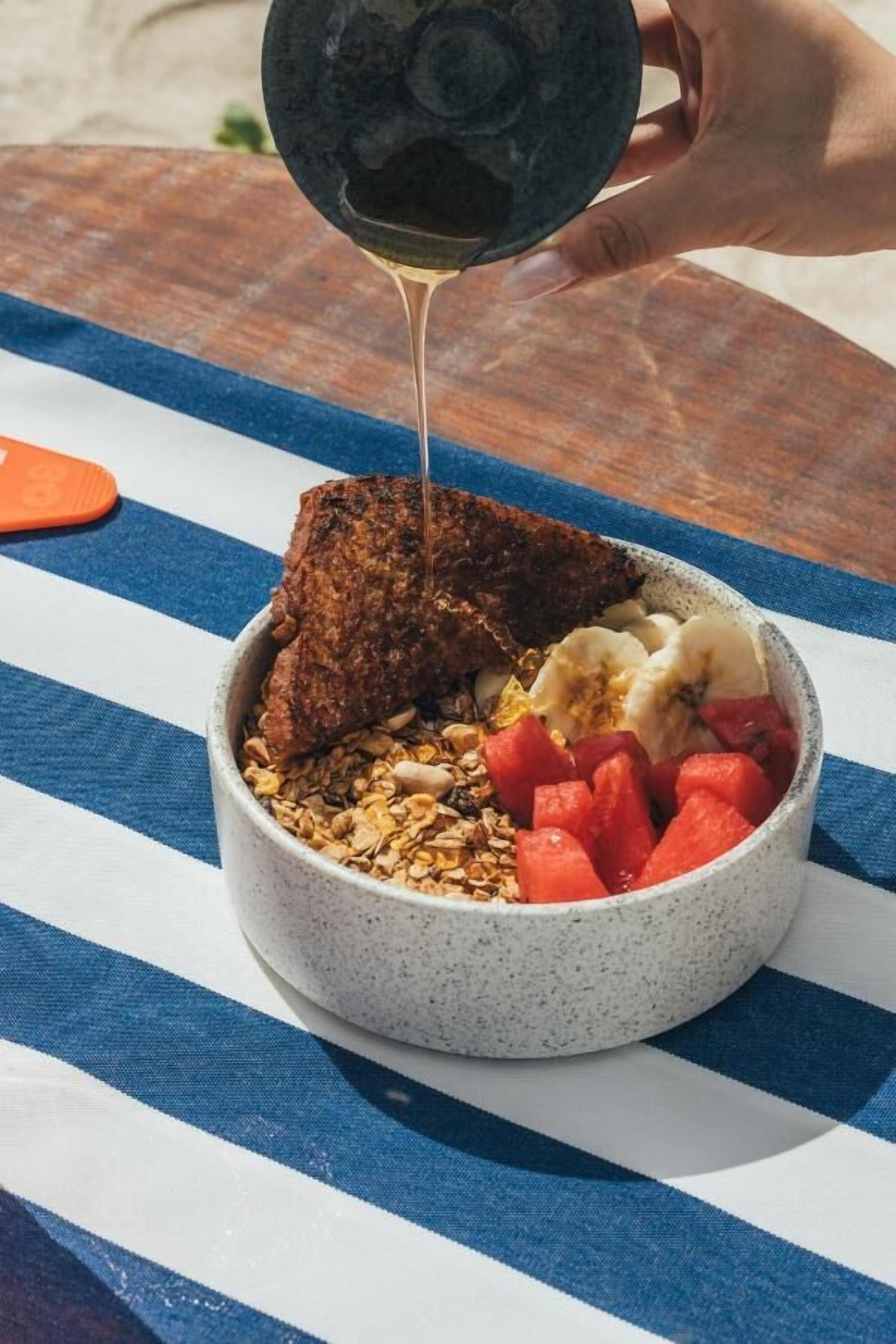 En skål med müsli, banan, vattenmelon och en bit rostat bröd, placerat på ett blå- och vitrandigt bordsduk.