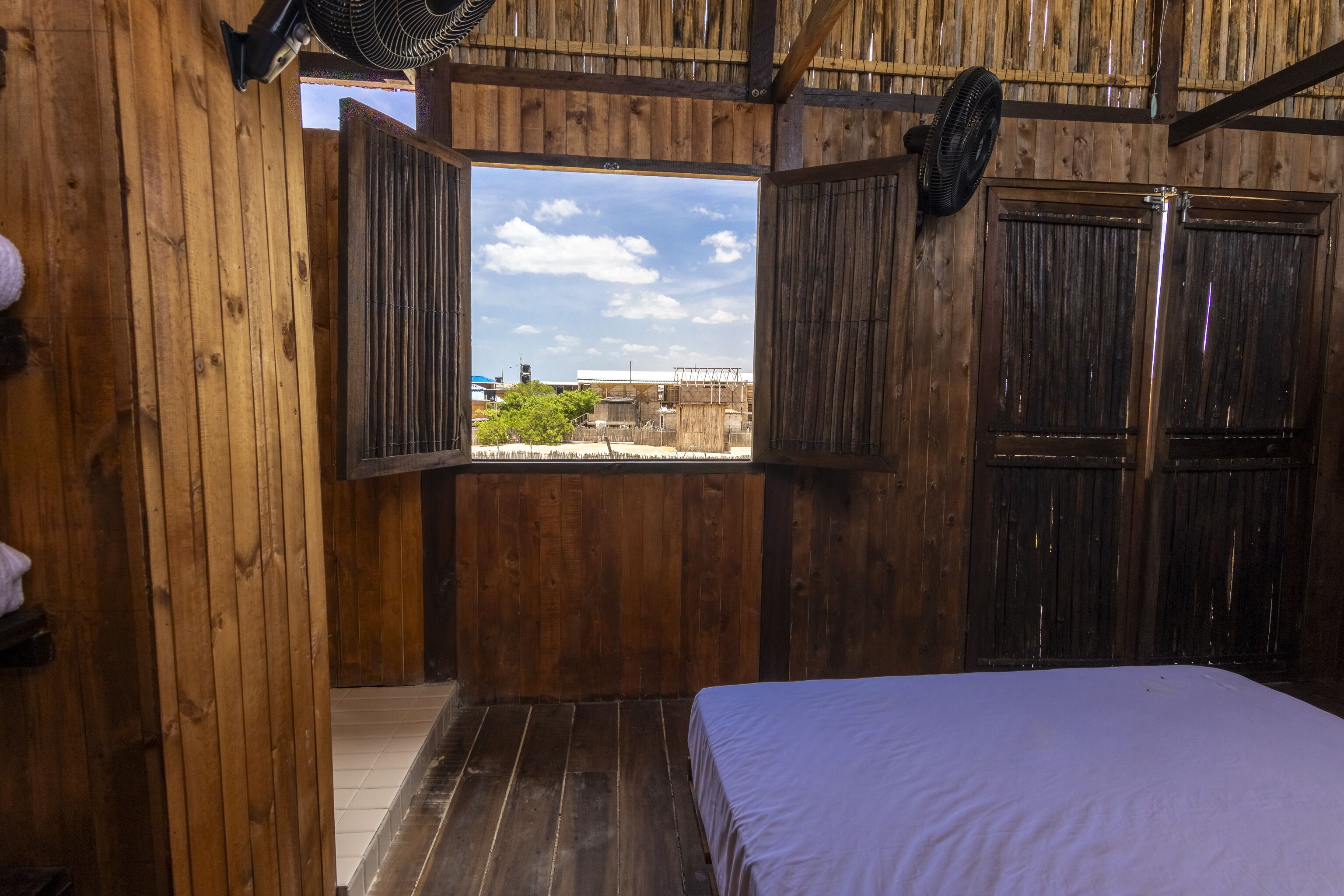 Ein Zimmer mit Holzboden, einem Bett, einem Fenster und einem Ventilator.