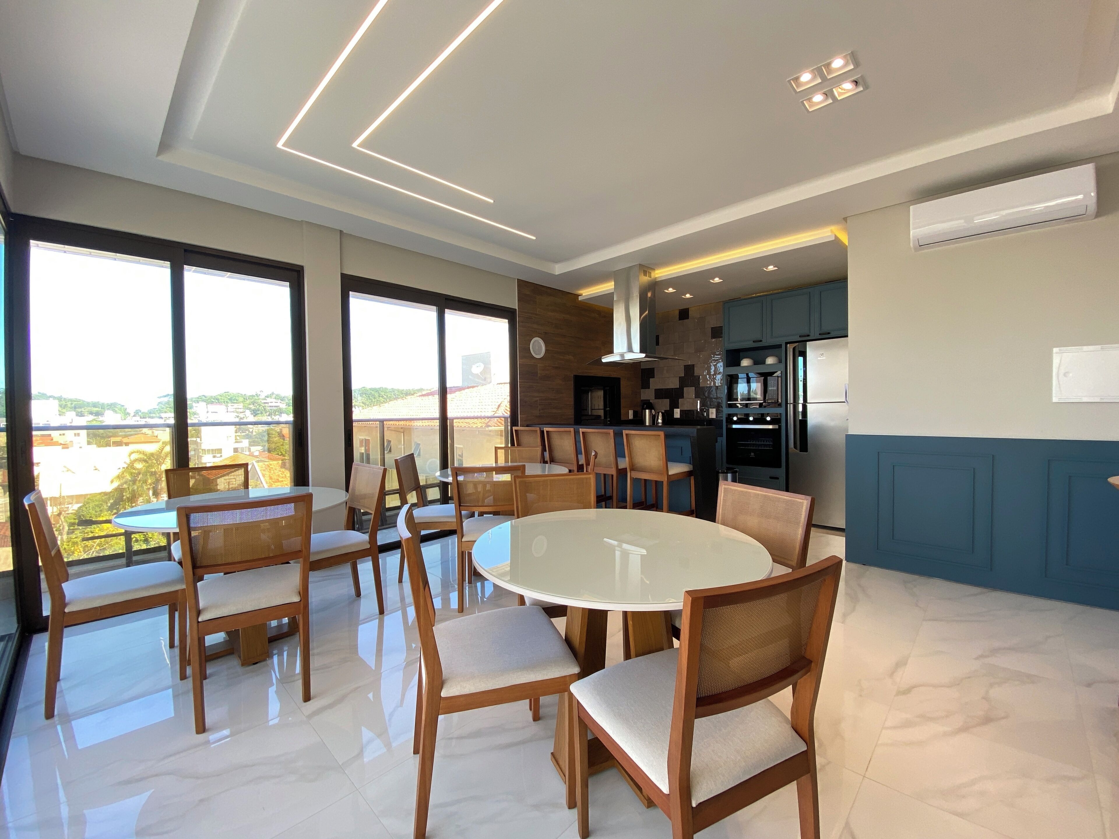 A modern dining area with a round table and chairs, large windows offering a view, and a kitchen area with blue cabinets and a refrigerator.