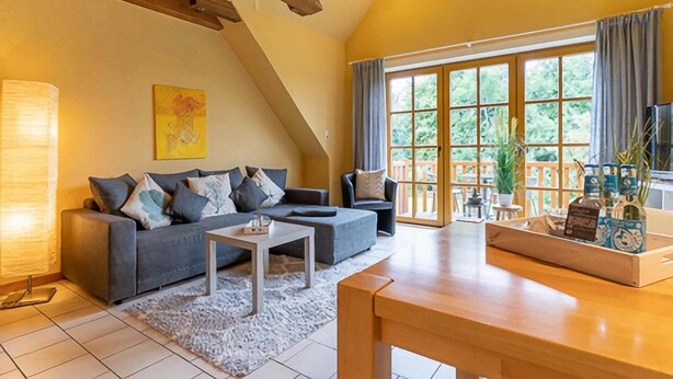 A living room with a grey sofa, a white coffee table, and a wooden dining table. There is a large window with curtains, a painting on the wall, and a lamp. - Landhaus in Vollerwiek (Vollerwiek)