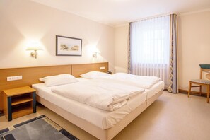 A hotel room with two beds, a small table, a chair, and a window with curtains.