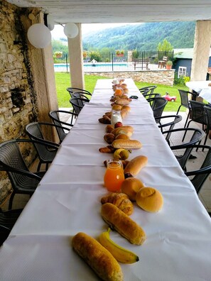 A long table with bread, a bottle of orange juice, and a banana, set up outdoors with chairs and a view of a pool and mountains in the background. - Beautiful Wedding venue for a destination wedding in Italy with pool, 32-50 pers (Bonvicino)