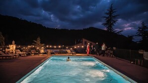 Ein Schwimmbecken mit Liegestühlen und einer Person, die schwimmt, umgeben von einem Zaun und Bäumen, unter einem bewölkten Himmel.
