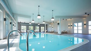 Una piscina interior con agua cristalina, rodeada de sillones de descanso y tumbonas, con luz natural entrando por amplias ventanas.