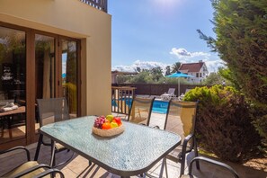 A glass-top table with a fruit basket, outdoor chairs, a pool, and a view of a house in the background.