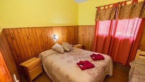 Une chambre avec une tête de lit en bois lambrissé, un lit recouvert d’une couvre-lit beige à motifs, deux oreillers et deux serviettes rouges. On y trouve une table de chevet en bois avec une lampe, une commode en bois et une fenêtre avec des rideaux rouges.