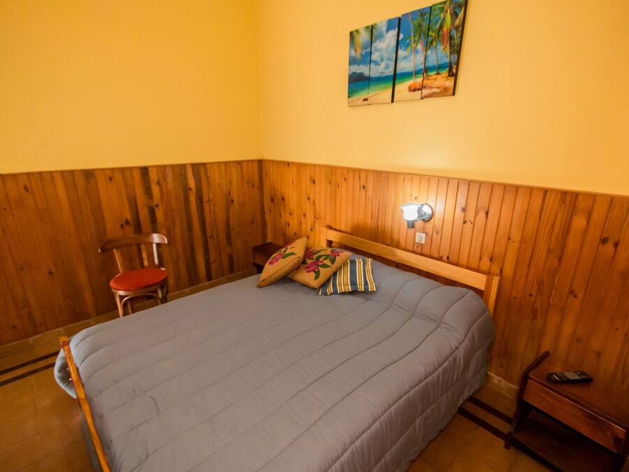 A bedroom with a wooden headboard, a bed with a grey bedspread, two pillows, a nightstand with a lamp, and a chair with a red cushion.
