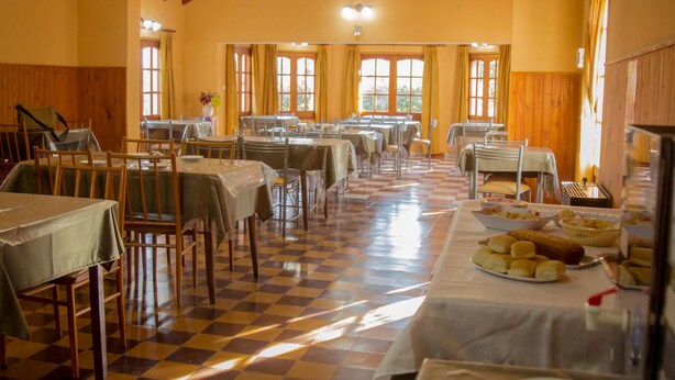 Un comedor con varias mesas y sillas, piso a cuadros y ventanas amplias con cortinas.