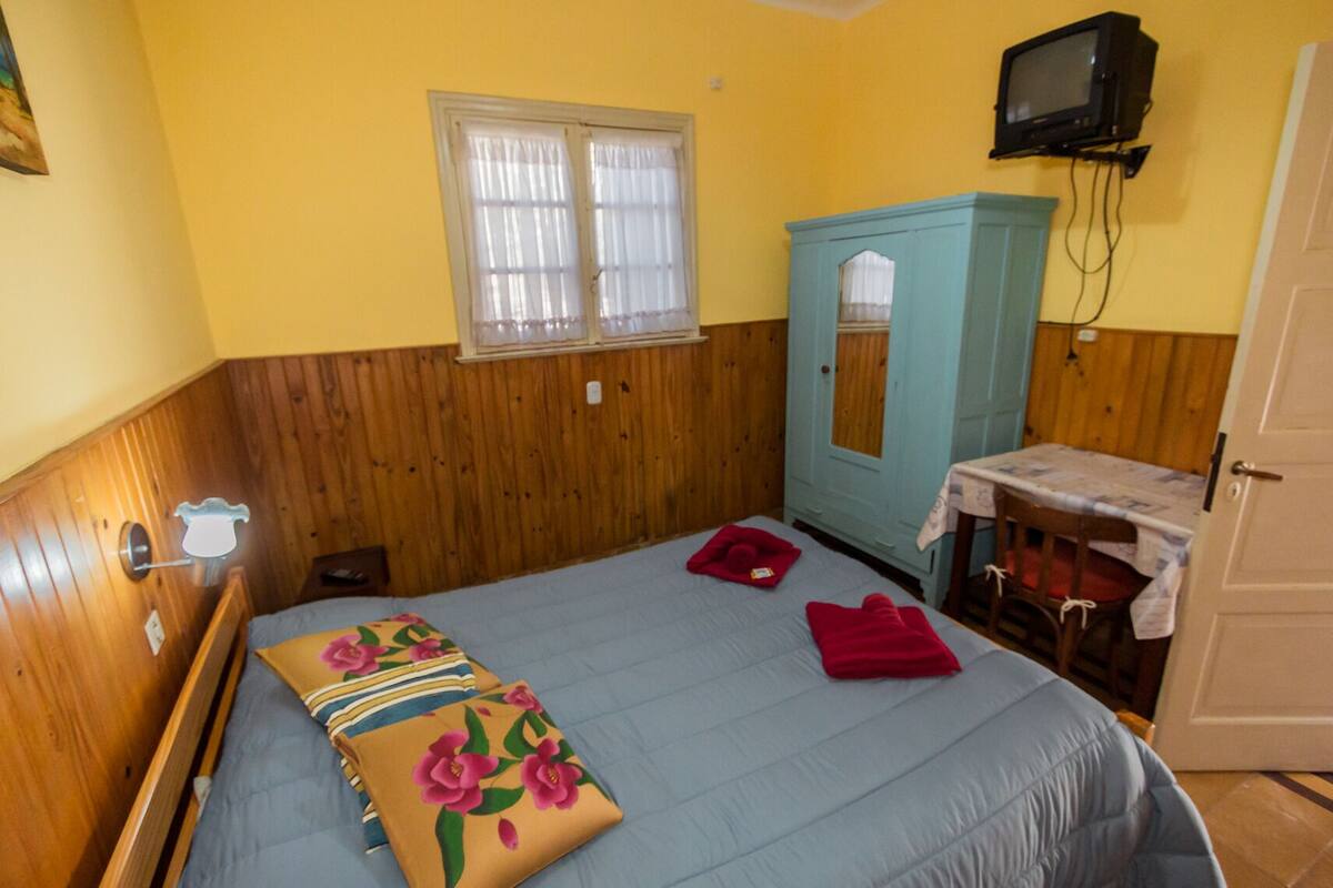 Une pièce comprenant un lit, une armoire en bois, une petite table recouverte d’une nappe, un téléviseur fixé au mur et une fenêtre avec des rideaux.
