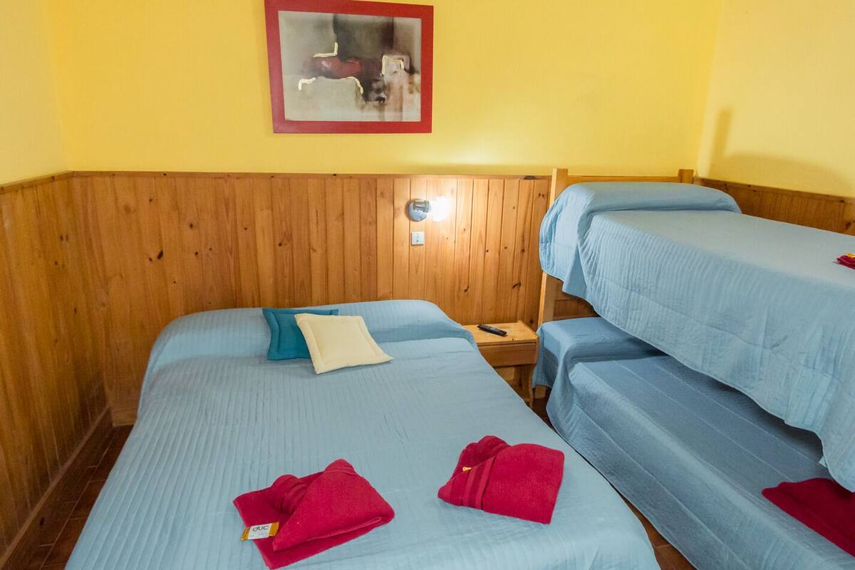 Une chambre avec deux lits, chacun recouvert d’une couverture bleue et agrémenté d’oreillers, une tête de lit en bois, une petite table de chevet avec une lampe, et un tableau encadré accroché au mur.