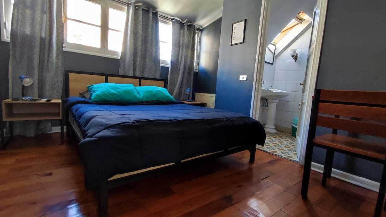 A room with a bed, a wooden chair, a nightstand, a window with curtains, and a bathroom with a sink and mirror.