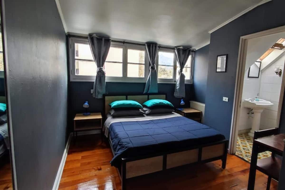 A bedroom with a bed, bedside tables, a window with curtains, a small table, and a bathroom with a sink and mirror.
