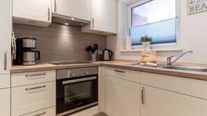 A modern kitchen with white cabinets, a built-in oven, a stainless steel coffee maker, and a window with a view of a brick building. - Ferienwohnung in St. Peter-Ording (St. Peter-Ording)