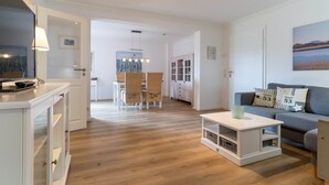 A modern living room with a grey sofa, a white coffee table, and a dining area with wicker chairs. - Ferienwohnung in St. Peter-Ording (St. Peter-Ording)