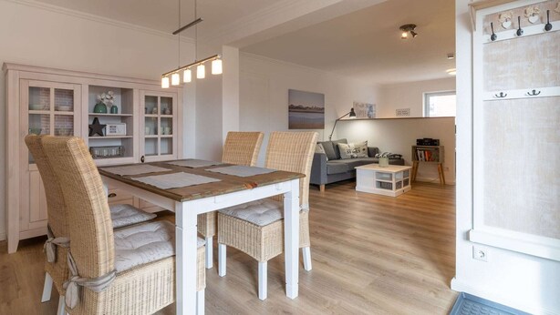 A dining area with a white table and wicker chairs, a kitchen with open shelving, and a living area with a sofa and a lamp. - Ferienwohnung in St. Peter-Ording (St. Peter-Ording)