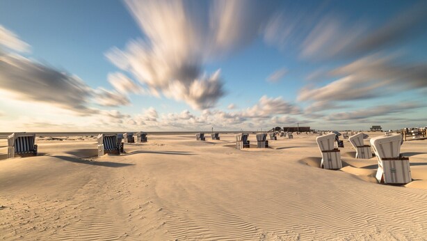 A beach with rows of white beach chairs and a few blue chairs, with a clear sky and clouds.